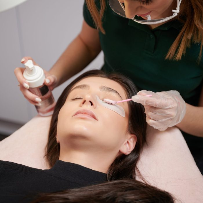 Close up of beauty specialist in sterile gloves applying cleansing mousse on woman eyelashes with cosmetic lash brush. Eyelash stylist preparing woman lashes for applying extensions.